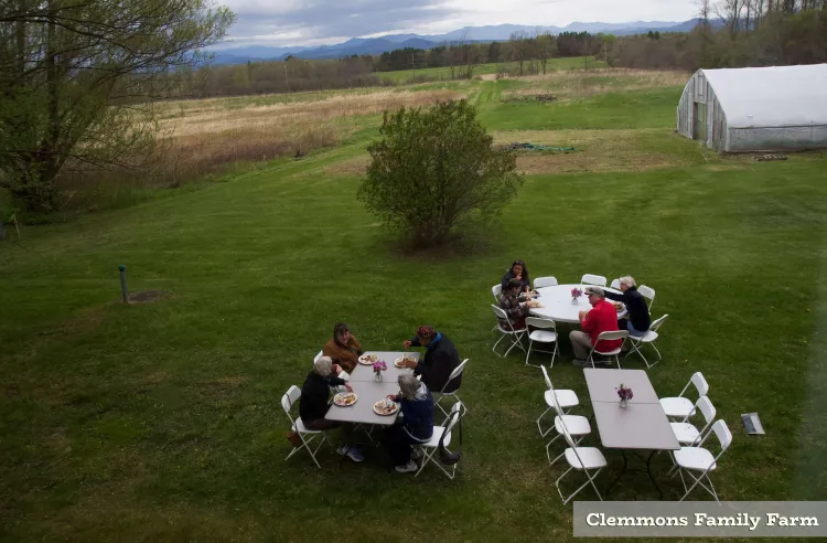 A view from above of three tables, two of which have people sitting around them, outside at a farm. The scene is grassy but early spring, so there is barely new growth on the trees and bushes in the background. Layers of blue mountains are visible in the distance, and there's a greenhouse off to the side.