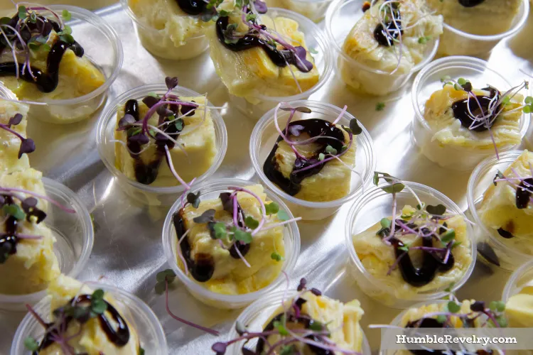 Small plastic cups with squares of a baked, yellow mac and cheese, topped with a brown glaze and a little greenery.