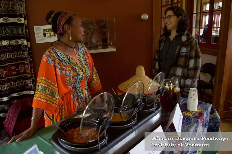 A Black woman in a bright orange, patterned dress stands in front of crockpots while talking with a white woman.