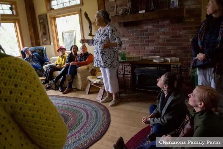 A Black woman speaks in front of a brick fireplace in a wood-floored, warm and bright room full of listening onlookers.
