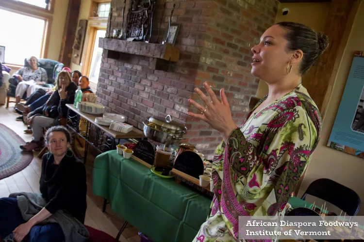 A biracial woman speaks and gestures with her hand in a green and pink jumpsuit in front of a roomful of people.