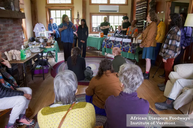 A roomful of people in a warm, bright space with windows, wooden floors and a brick fireplace, with everyone's attention directed at a Black woman speaking in the center. Behind her are tables with food laid out.