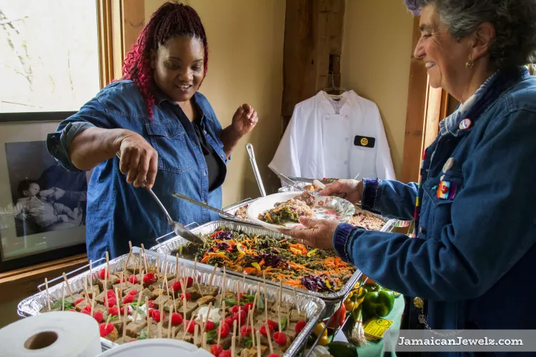 A Black woman smiles slightly while serving brightly colored salad from a large tray to an also-smiling tanned-skin woman holding a plate.