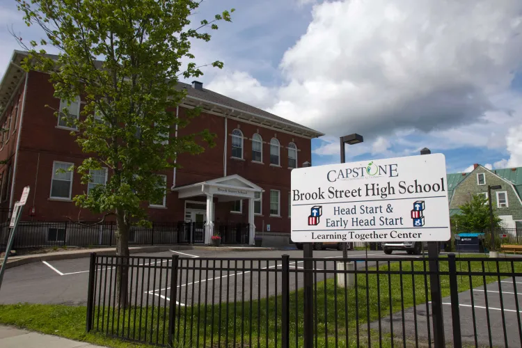 A white sign reading "Capstone community action Brook Street High School head start and early head start learning together center" is outside a brick building on a partly-cloudy day in summertime.
