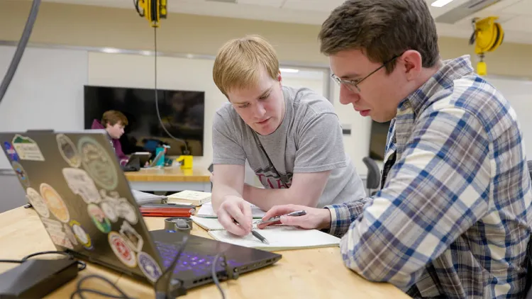 Two students discussing homework at a table with an open laptop