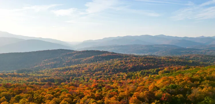 Photograph of a mountainous landscape in Vermont during autumn showcasing vibrant fall foliage in shades of orange, red, and green across rolling hills. Distant mountain ranges under a partly cloudy sky create depth and highlight seasonal transition.