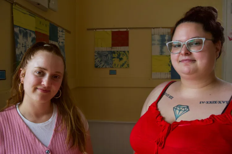 Two white women, one in a red tank top and glasses, the other in a pink sweater and golden hoops, stand side by side with relaxed looks on their faces. They're lit up with natural light in a yellow-painted room with quilt squares behind them.