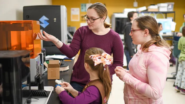 Two grade-school students are provided a demonstration of lab equipment by an undergraduate lab technician.