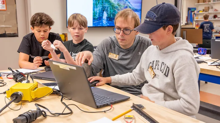 A camp instructor works with a student at a laptop while other students look on