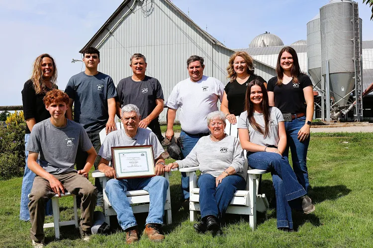 A posed portrait with ten people in front of a white barn and silos on a sunny, summer day. 