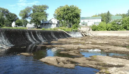 A photograph showing a view of the Swanton Village Dam with houses behind it on a sunny day in 2015