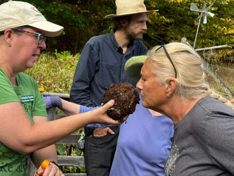 One white woman holds out a ball of soil while a second white woman sniffs it. A third person stands looking off to the side in the background. They're all in a garden environment.