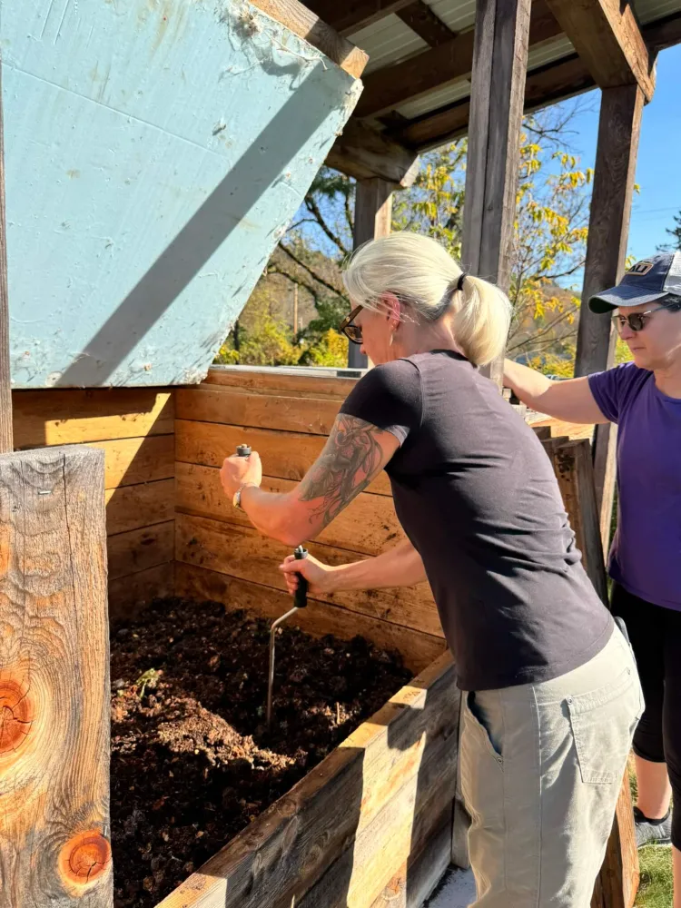 A white woman sticks a metal rod with a handle in a wooden bin full of soil.