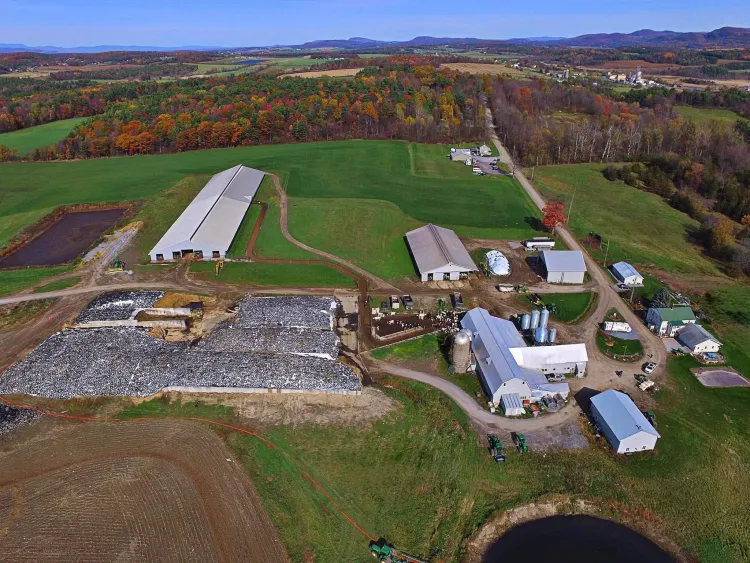 An overhead scene of a large farm with numerous buildings in the foreground and fields in the background, stretching into forested parcels of land and mountains in the furthest background. The trees are red and orange while the fields are bright green.