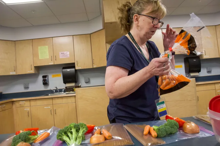 An adult white woman in front of pre-portioned pasta, broccoli, peppers and carrots in a kitchen with multiple work stations. She's holding a bag of carrots.
