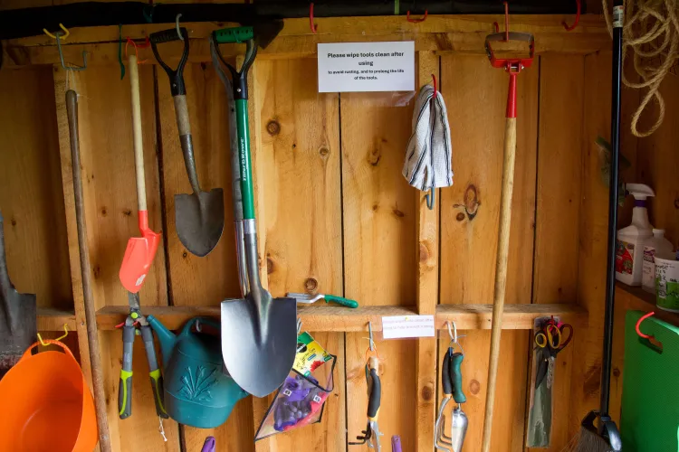 A wooden wall with many gardening tools hanging on it. A sign reads "please wipe tools after using to avoid rusting and to prolong the life of the tools."
