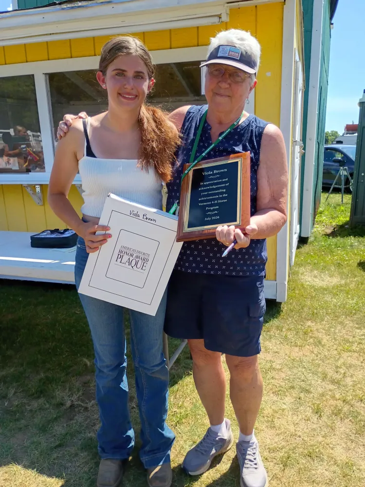 A white teen girl and an older white woman stand together outside a yellow building. The older woman holds a plaque reading: "Viola Brown: In appreciation and acknowledgement of your outstanding achievements in the Vermont 4-H Horse Program." The younger woman is holding a white box that reads "America's Favorite Honor Award Plaque."