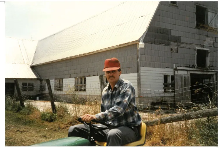 A scanned film photo showing a white man with a mustache in a bright red baseball hat and flannel in front of a large grey barn on a sunny day. He's riding on a lawn mower or a tractor. 
