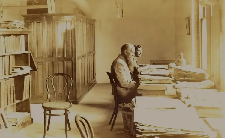Two men sit at a long table full of plant specimens, herbarium cabinet and bookshelf in background
