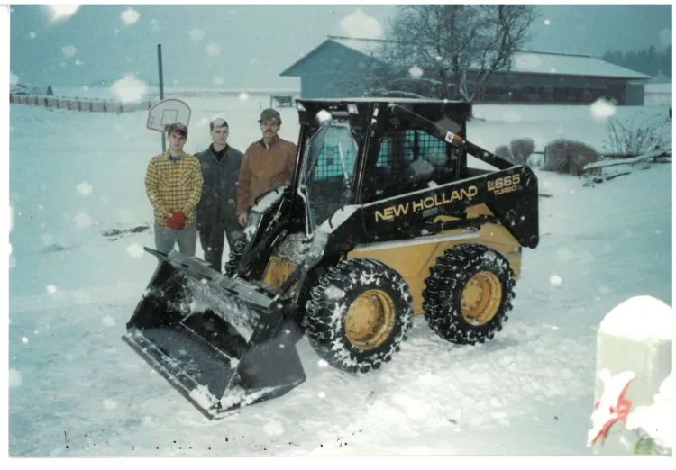 Two young white men and an older white man stand together in a snowy scene next to a small yellow and black tractor. A barn building is in the background, and snowflakes are illuminated with a flash in the foreground.