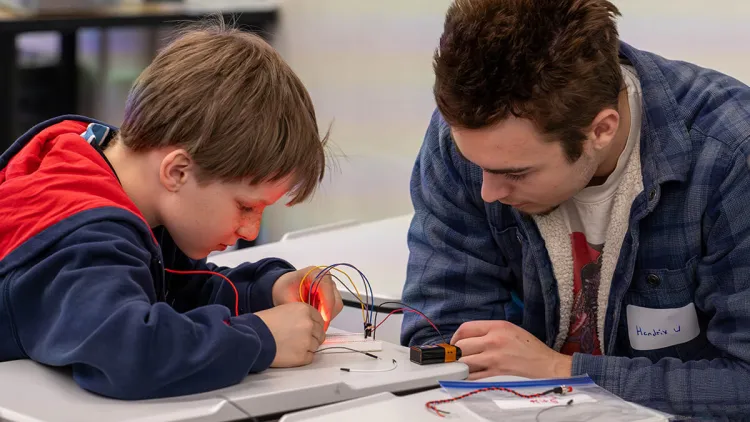 A college student helps a middle-school student complete an electrical circuit to light a small LED lamp