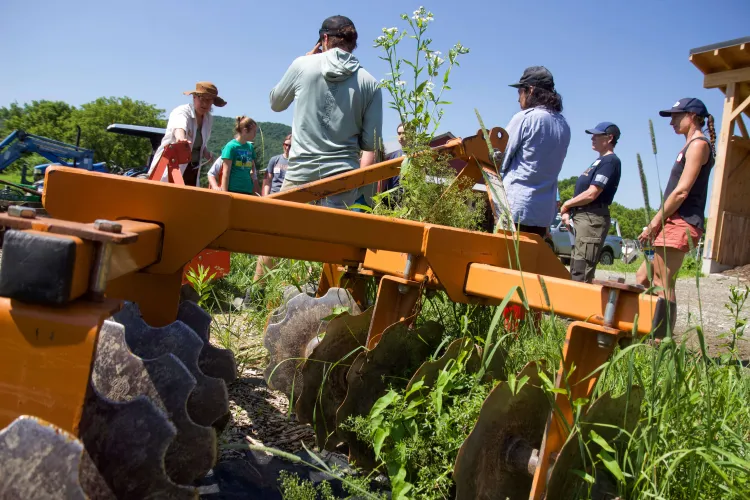 A group of people stand outside on a sunny day and are seen behind tractor hitch-on equipment that has large sharp disks. 