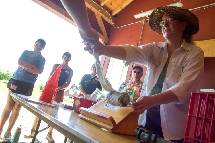 Inside a shed, a white woman in a white shirt and straw hat holds out a strip of fabric while more people look on..