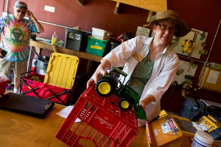 A white woman in a white top, straw hat and glasses tips a red crate with a toy tractor on top of it in a demonstration. 