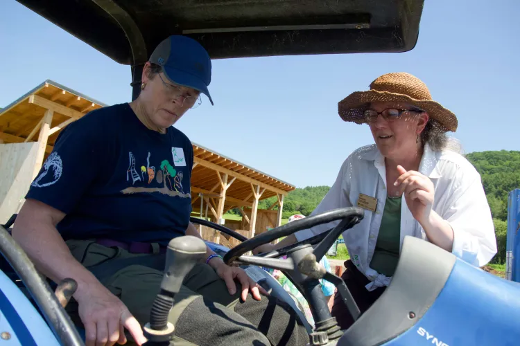 Two white femmes, one seated in a blue tractor, the second leaning over the tractor seat and pointing, are engaged in teaching and learning together.