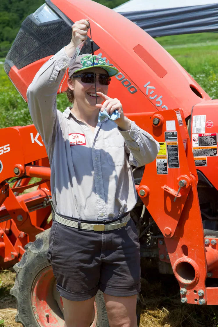 A femme white person wearing shorts, a button down shirt and blue-green patterned hat and sunglasses holds up a dipstick in front of an orange tractor.