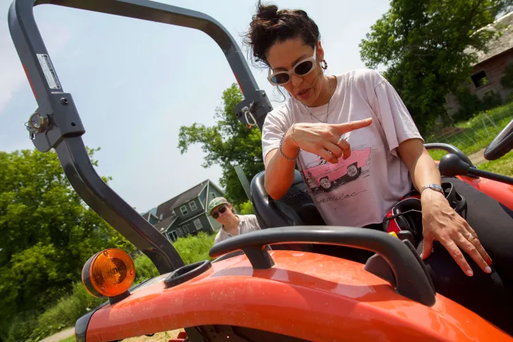 A close up of a brown-skinned woman with sunglasses and a pink shirt pointing while sitting on top of an orange tractor, while a femme person with a hat and sunglasses stands in the background looking on.