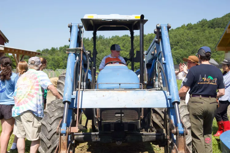 A preteen boy sits in a bright blue bucket, seen from the front of the tractor, and had his head turned while he listens to an adult. More adults and young people surround the tractor. It's a bright, blue-sky day and greenery is in the background.