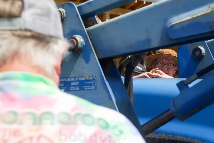 A close-up view of a white woman in a straw hat and glasses demonstrating with her hands, holding her fingers interlocked. She's seen through the space between a tractor's hood and the bucket mechanism. In the foreground is an onlooking person.