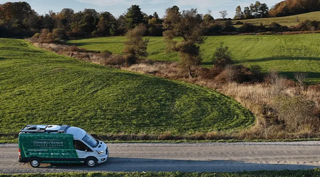 A drone shot of the UVM Cancer Center van out in the countryside driving along a dirt road. 