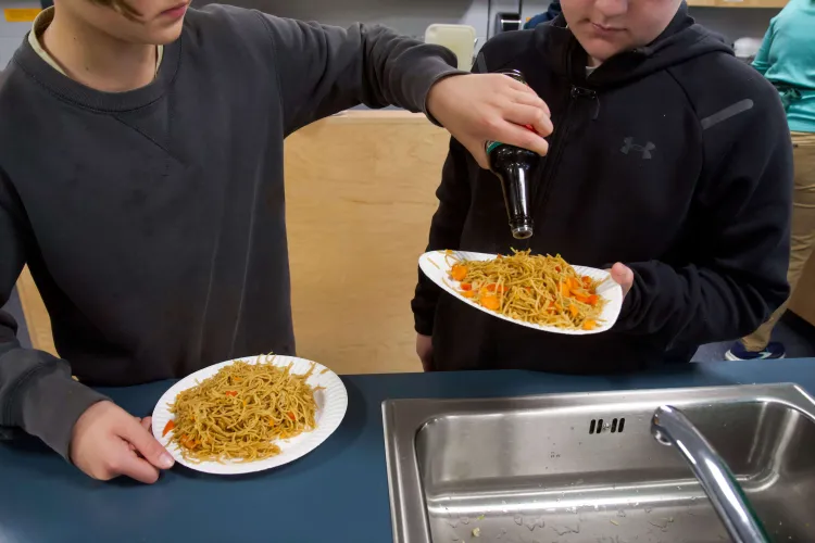 Two young people in sweatshirts, seen from the nose down, holding paper plates of veggie lo mein in a kitchen. One of the kids is pouring soy sauce on the other kid's plate.