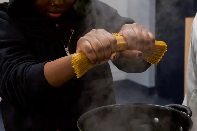 A Black child in a sweatshirt, seen from the nose down, holds a large bunch of dried spaghetti and bends it over a steaming pot.