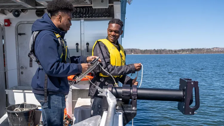 Two students lower a water monitoring device from the deck of a science vessel on Lake Champlain