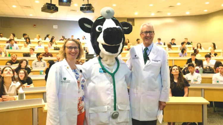 two doctors stand arm-in-arm with a cow as a doctor mascot, all in white coats