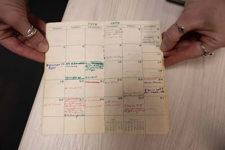Hands are holding open an old planner displaying a July 1976 spread. Throughout the page are notes written in different pen colors.