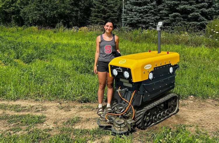 A young woman with dark brown hair in a ponytail, in a grey tank stop and black shorts, stands next to a yellow, lawnmower-sized robot in a field. 