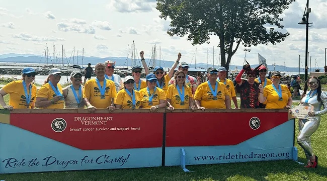 Group of people in yellow shirts with blue medals hanging around their necks outside cheering. In front of them is a banner that reads "Be the Change" for the 2026 Dragon boat race. 