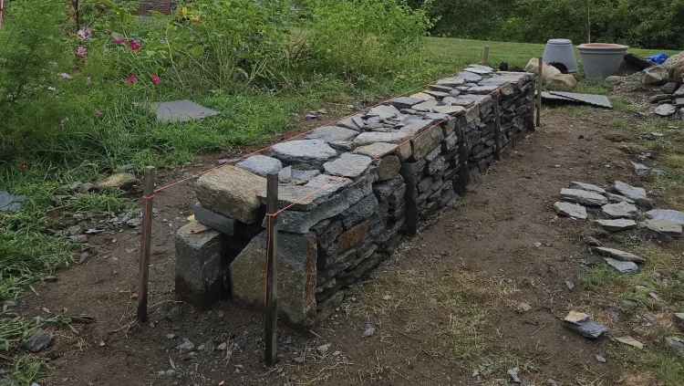 A dry stone wall made of slabs of rock stacked into a rectangular shape. It sits along the edge of a garden. 