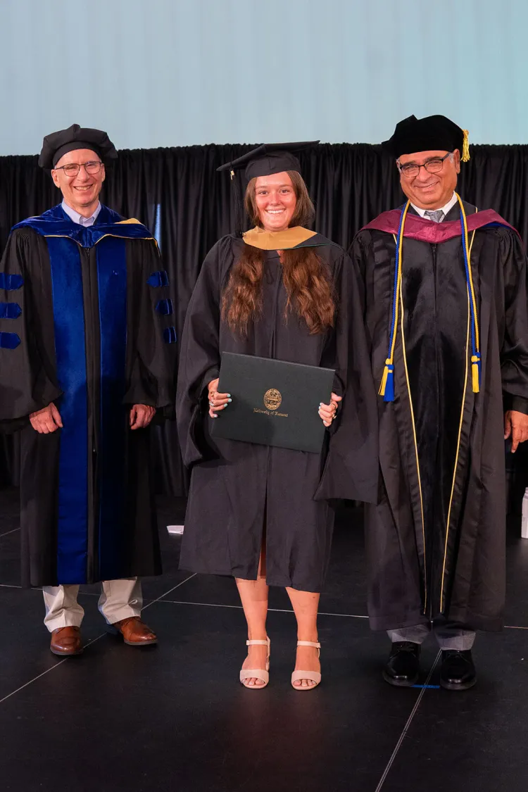 A graduating SI-MBA student in cap and gown holds their diploma on stage, flanked by Dean Berger and Dean Anand in their regalia