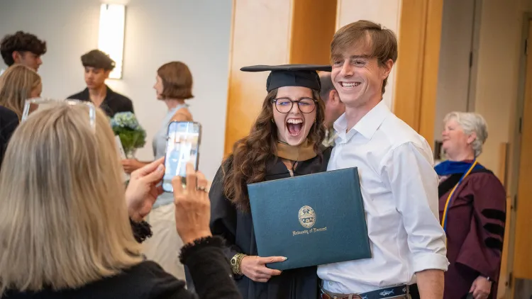 A graduating SI-MBA student in cap and gown and holding their diploma shares their elation with family