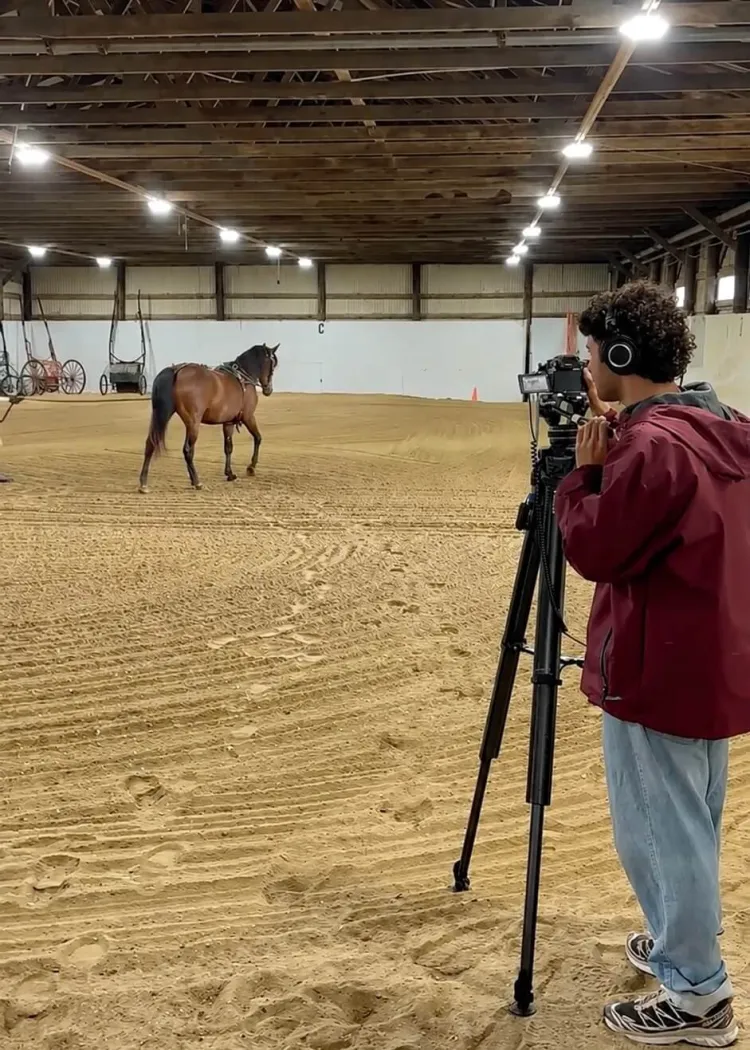 A young person with short brown curly hair wearing a maroon jacket, jeans and headphones stands behind a video camera in a riding ring where a horse is walking in the distance.