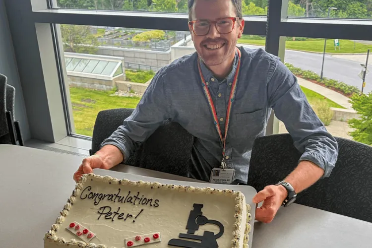 Person in a blue button up, a rainbow lanyard, and tortoise glasses is holidng a cake that says "Congratulations Peter!" in front of a window that overlooks a road. 