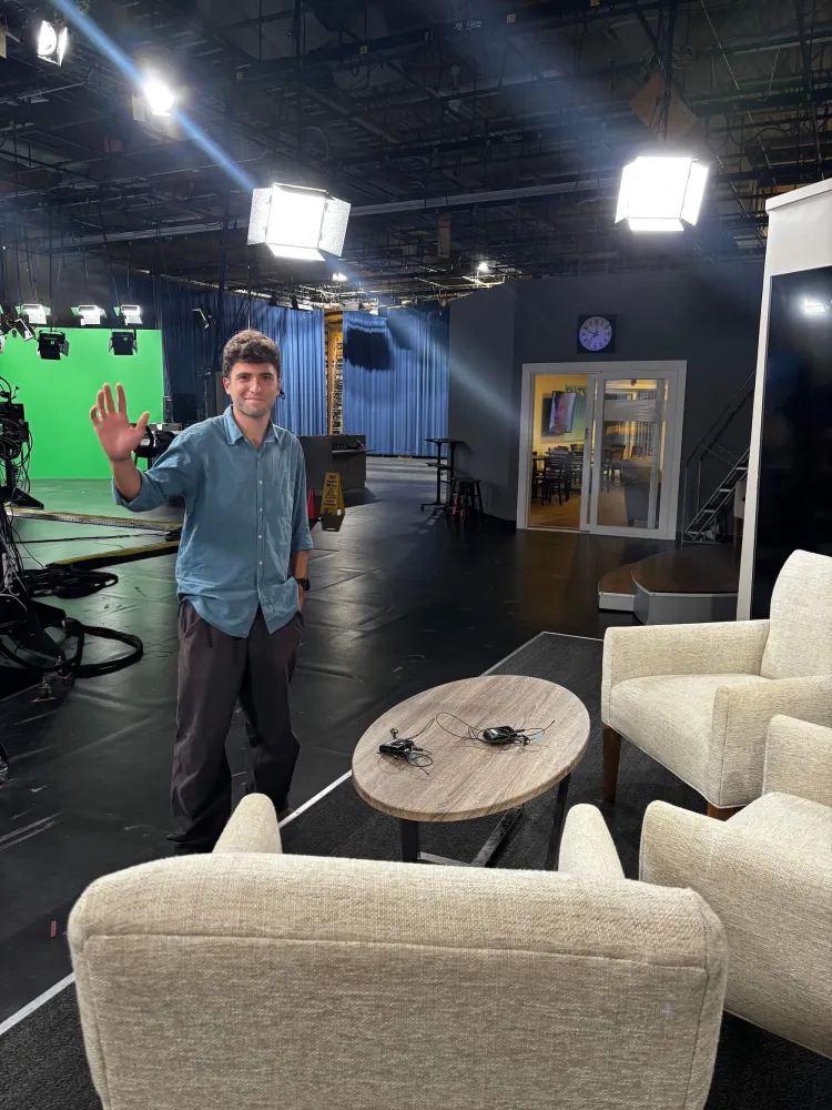 A young man with short curly brown hair waves inside a TV studio where there are comfy chairs, a small round table and bright overhead lights. 