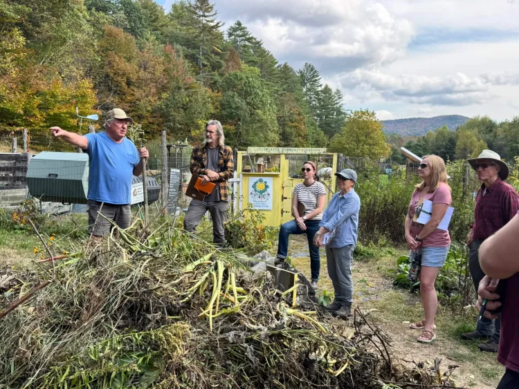 A group of six people stand outside in front of a pile of yard scraps while a white man in a sky-blue t-shirt and baseball hat points and speaks.