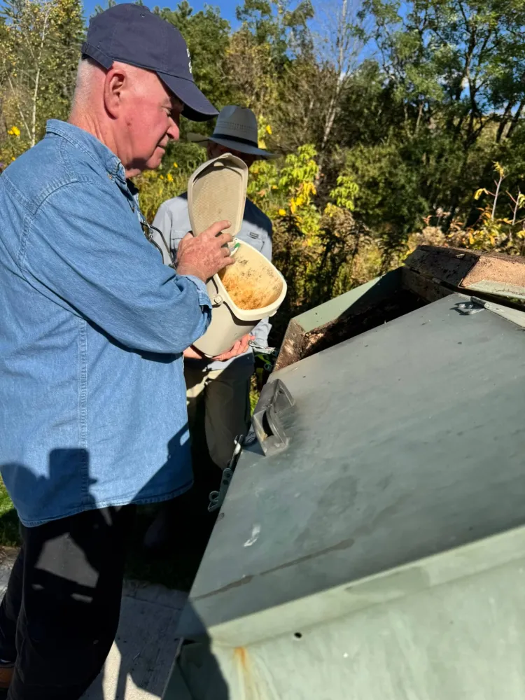 A white man in a blue baseball hat and denim long-sleeved shirt holds a bucket full of food scraps over a larger bin. 