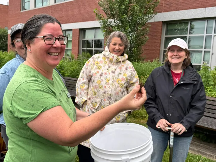 Four people smile and look at the camera outside a brick building. The person in the foreground is holding a white bucket and a small piece of soil up.